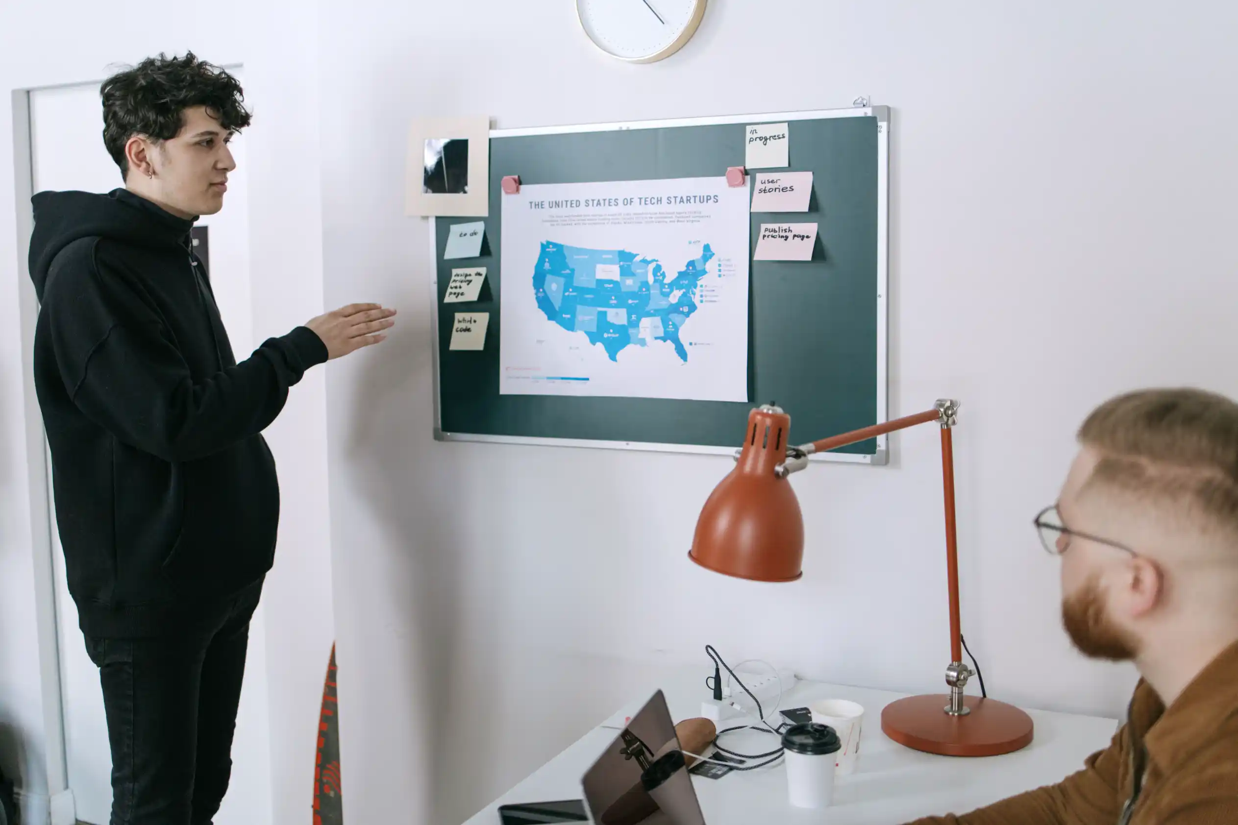 A man pointing at a map on an office board while a colleague takes notes