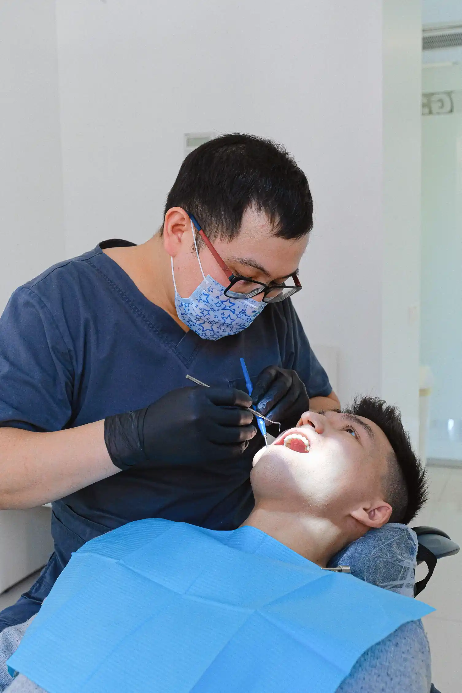 A dentist in glasses and navy scrubs examining a patient's teeth
