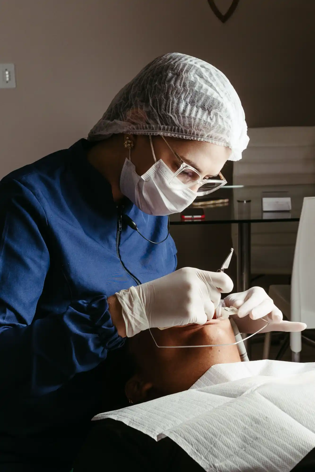 A dental clinician in a mask and hair cover treating a patient